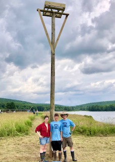 Noah stands at the base of a tall osprey nesting pole. Two team members stand at his side.
